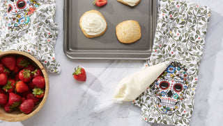 A bowl of strawberries, a baking tray with cookies, a piping bag of icing, and two skull-patterned towels are arranged on a marble countertop.