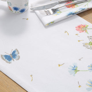 A white tablecloth with floral and butterfly designs, a matching napkin with cutlery on top, and part of a floral-patterned cup, all on a light wooden table.