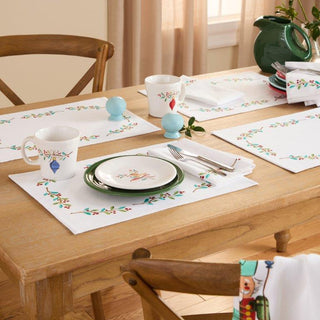 A wooden dining table set with bird-themed placemats, plates, and mugs, green teapot, napkins, cutlery, and salt and pepper shakers. Sunlight comes through a nearby window.