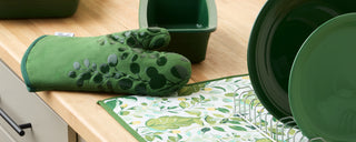 A green oven mitt with a leaf pattern rests on a wooden counter next to green dishes, a drying rack, and a dish towel with a leafy design.