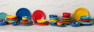 A colorful collection of ceramic plates, bowls, and dishes in red, yellow, blue, and turquoise is neatly arranged on a white marble surface against a matching marble background.