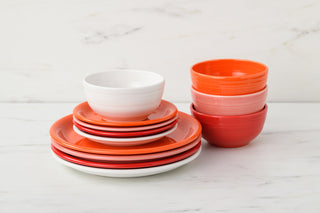 A stack of red, orange, and white plates and bowls sits on a white marble surface with a light background. The dishes are neatly arranged in color-coordinated stacks.