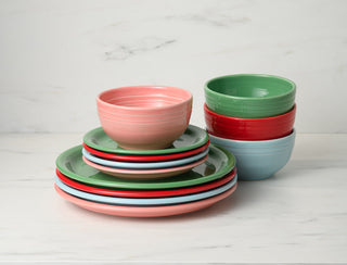 Stacks of colorful ceramic bowls and plates in pastel pink, green, red, and blue sit on a white marble surface with a white marbled background.