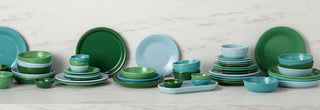 Assorted green, blue, and teal ceramic dishes, bowls, and plates are stacked and arranged on a white marble surface against a marble-patterned background.