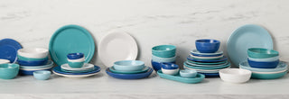 A variety of stacked plates, bowls, and cups in shades of blue, teal, and white are arranged on a white marble countertop with a marble backsplash in the background.