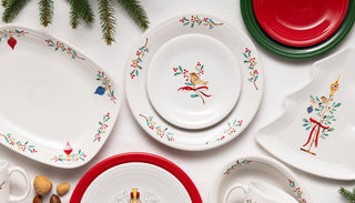 A festive table setting with white plates decorated with birds, holly, and red ribbons, alongside red and green plates. Evergreen branches and assorted nuts accent the holiday theme.
