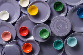 Assorted purple plates with embossed rings are arranged with colorful bowls in orange, yellow, green, red, peach, teal, and white. A purple pitcher and a plate labeled Fiesta are included.