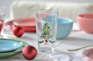 A clear glass with a colorful Christmas tree design sits on a festive table set with pastel plates, bowls, and red ornaments, creating a cheerful holiday atmosphere.
