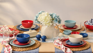 A dining table set with red, blue, and cream dishes on woven placemats, glassware, napkins, and a white pitcher with white and blue flowers as a centerpiece. The table has a casual, inviting feel.