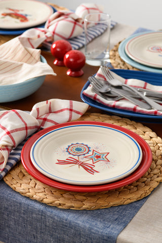 A festive table setting with woven placemats, patriotic-themed plates, red and white napkins, utensils, glasses, and red salt and pepper shakers on a wooden table.