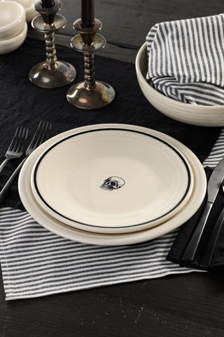 Four cream-colored plates with black rims, each featuring a different Halloween-themed design: a raven, a witch hat, a spider web, and a skull, arranged in a row.