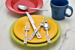 A colorful table setting with a green plate under a yellow plate, silverware with multicolored striped handles, a red bowl with a spoon, and a blue mug on a marble surface.