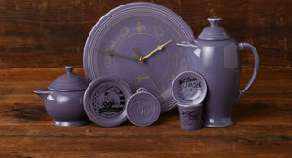 Lavender ceramic teapot, sugar bowl, and clock with gold hands, plus commemorative round containers, all arranged on a wooden surface with a wood background.