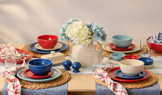 A wooden table set for four with red, blue, and cream dishes, woven placemats, glasses, napkins, and a white pitcher holding blue and white flowers as the centerpiece.