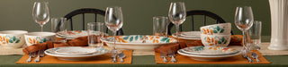 A dining table set for two with autumn-themed plates, bowls, glasses, and brown napkins on orange placemats, against a green background with dark wooden chairs.