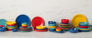 A variety of colorful dishes, bowls, and plates in red, yellow, blue, and teal are neatly stacked on a white marble countertop against a white marble background.