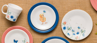 Assorted ceramic plates and a mug with blue and pink rims, featuring floral designs, a chick, and a butterfly, arranged on a burlap surface.
