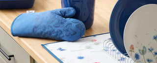 A blue oven mitt with butterfly designs rests on a wooden countertop next to a dish rack holding blue and floral-patterned plates and a blue ceramic container.