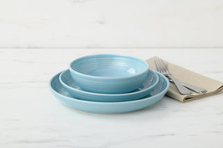 Three stacked light blue bowls from the Fiesta Coupe Bowl 3-Piece Dinnerware Set, Service for 1, sit on a white marble surface next to a folded beige napkin with a fork and knife placed on top.