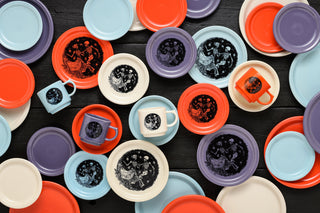 Colorful plates with skull and skeleton couple designs are surrounded by vibrant red, yellow, and purple flowers arranged on a purple background, creating a festive and lively display.