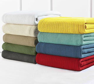 A stack of nine neatly folded knitted blankets in various colors, including white, yellow, cream, beige, green, blue, teal, gray, and red, arranged on a white surface against a white wall.