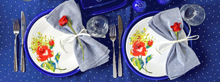 Two place settings with blue and white floral plates, gray napkins tied with ribbon and red roses, clear glasses, silverware, and a blue tablecloth with white polka dots.