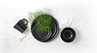 Black ceramic plates, a mug, and a bowl are arranged on a white wooden surface with a white napkin and a green fern, creating a minimalist, modern table setting.