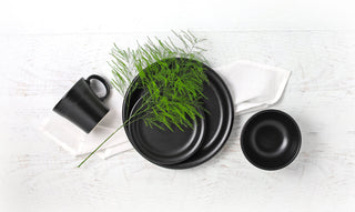 Black ceramic plates, a bowl, and a mug are arranged on a white cloth with a green leafy sprig on top, set on a white wooden surface.