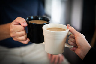 Two people clink a black mug and a white mug filled with coffee, with blurred background suggesting a casual, friendly setting.