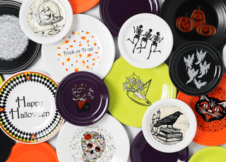 An assortment of colorful Halloween-themed plates featuring designs like skeletons, pumpkins, witches, cats, skulls, and festive phrases, arranged overlapping on a white surface.