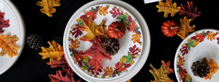 Three white plates with autumn leaf borders, each decorated with pinecones, faux leaves, and small pumpkins on a black surface, surrounded by scattered fall leaves.