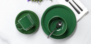 A green cup, saucer, bowl, and two plates are arranged on a white dotted surface with a spoon, black cutlery on a napkin, and some purple flowers in the corner.