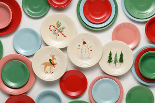 Assorted red, green, blue, and pink plates and bowls, with three white plates featuring Christmas designs: a reindeer, a candy cane with pine, and two decorated Christmas trees.