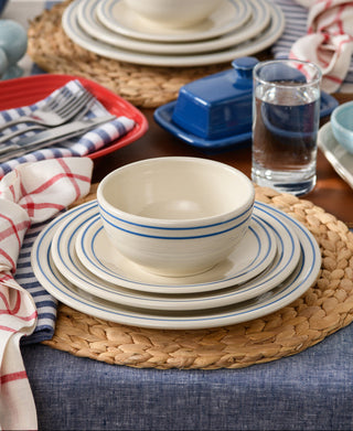 A place setting with stacked cream dishes featuring blue stripes, a bowl on top, woven placemat, checkered napkin, glass of water, blue butter dish, and a red dish on a wooden table with blue tablecloth.