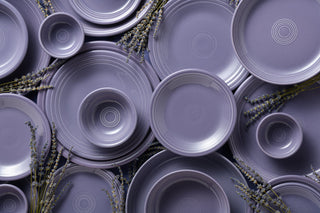 A dining table set with lavender-colored dishes, glassware with white wine, napkins, silverware, and dried lavender. The table has a woven runner and is decorated with natural, rustic elements.