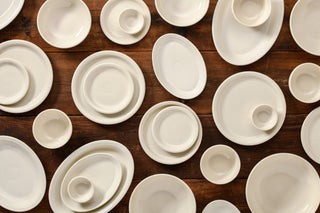 Assorted white ceramic plates, bowls, and cups arranged on a rustic wooden table, viewed from above.