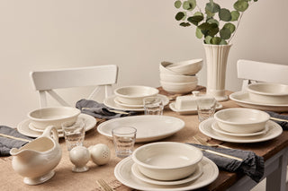 A dining table set with cream-colored ceramic dishes, glass cups, gray napkins, salt and pepper shakers, and a vase with green eucalyptus branches, arranged on a tan woven tablecloth.