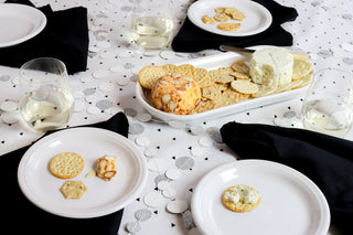 A table set with white plates, black napkins, wine glasses, crackers, and two cheese balls, one coated in herbs and the other with nuts. Crackers with cheese are on the plates.
