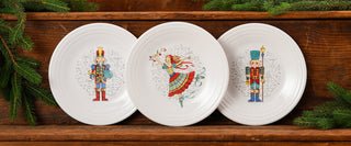 Three white plates with festive designs: a nutcracker on the left, a dancing ballerina in the center, and another nutcracker on the right, displayed on a wooden shelf with evergreen branches.