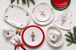 Festive ceramic dinnerware set with holiday designs, including a nutcracker, ornaments, birds, and holly, arranged with pine branches, nuts, and a napkin tied with a red ribbon.