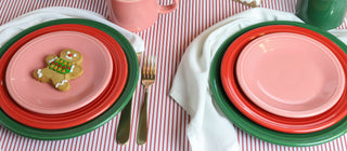 A festive table setting with green and red plates, pink mugs, white napkins, and a gingerbread cookie on one plate, arranged on a red and white striped tablecloth.