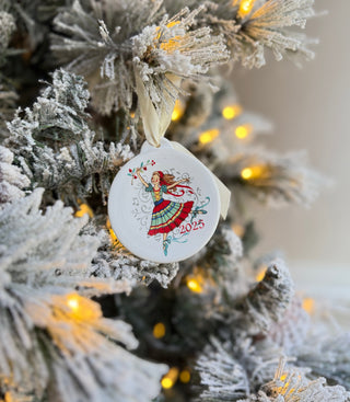 A decorated Christmas tree branch with snow-covered needles, glowing yellow lights, and a round ornament showing a dancing woman in festive clothes with 2025 written on it.