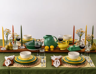 A colorful table setting with green, yellow, and cream dishes, glasses of wine and water, taper candles, pears, lemons, small flower vases, and napkins on woven placemats and an olive tablecloth.