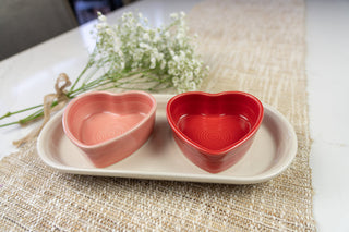 Two heart-shaped bowls, one pink and one red, sit on a white oval tray atop a woven table runner, with a small bouquet of white flowers in the background.