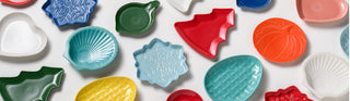 Colorful ceramic dishes in various shapes, including hearts, trees, shells, snowflakes, and pumpkins, are scattered on a white background. The dishes feature festive and seasonal designs.