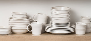 A collection of white ceramic bowls, plates, mugs, and cups neatly stacked and arranged on a wooden shelf against a plain white wall.