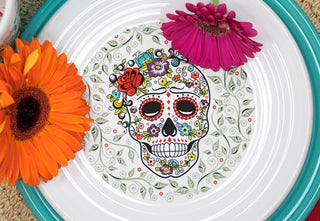 A decorative plate with a colorful sugar skull design surrounded by floral patterns, topped with an orange and a magenta daisy flower on the rim.