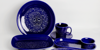 A set of deep blue ceramic dishes featuring intricate white skull designs, including plates, trays, a cup with saucer, and a small bowl, arranged on a white surface.