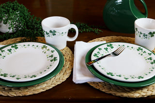 Two place settings with plates, cups, and bowls decorated with green shamrocks sit on woven placemats. A white napkin with a fork is on one plate. Greenery and a green pitcher are in the background.
