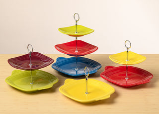 Three colorful tiered dessert stands with red, yellow, and blue square plates sit on a wooden surface, each with a silver ring handle on top.